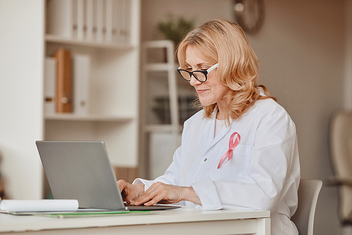 Warm-toned portrait of mature female doctor with pink ribbon pinned on white lab coat while working at laptop in modern office, breast cancer awareness symbol, copy space