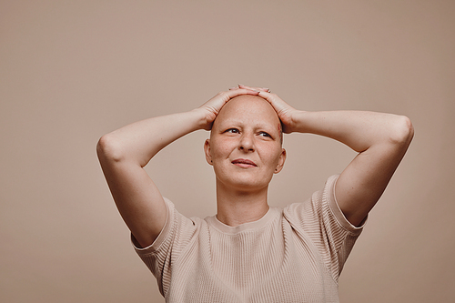 Warm-toned waist up portrait of confident bald woman looking away with hands on head while posing against minimal beige background in studio, alopecia and cancer awareness, copy space