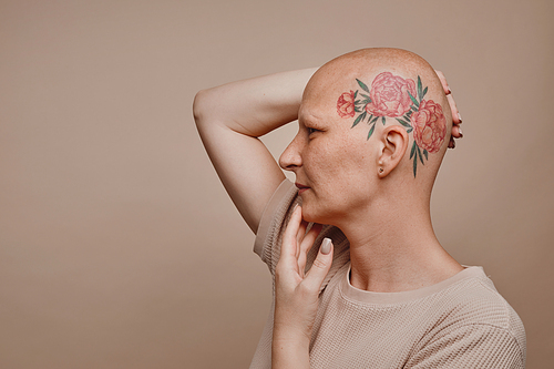 Minimal side view portrait of bald woman with head tattoo posing against beige background in studio, alopecia and cancer awareness, copy space
