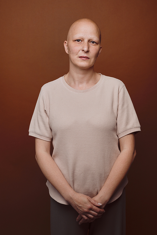 Minimal waist up portrait of bald woman looking at camera while posing against plain background in studio, alopecia and cancer awareness