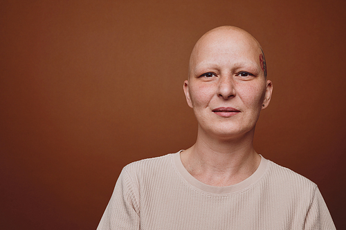 Minimal head and shoulders portrait of bald woman smiling at camera while posing against warm toned background in studio, alopecia and cancer awareness, copy space