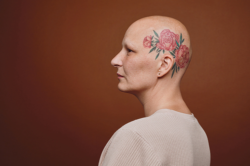 Minimal side view portrait of confident bald woman with head tattoo posing against brown background in studio, alopecia and cancer awareness, copy space