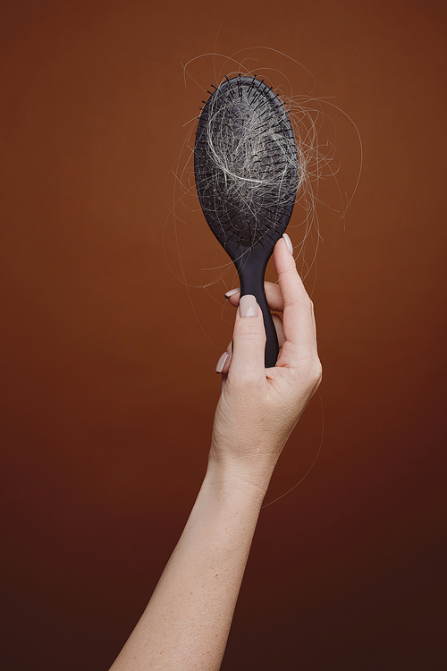Vertical background image of female hand holding brush full of hair against brown background in studio, alopecia and hair loss concept, copy space