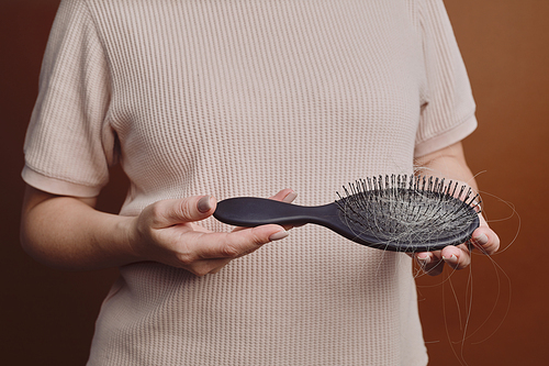 Cropped image of unrecognizable woman holding brush full of hair against brown background in studio, alopecia and hair loss concept, copy space