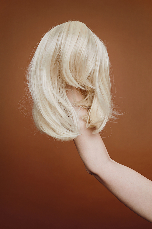 Vertical background image of female hand holding wig of blonde hair against brown background in studio, alopecia and hair loss concept, copy space