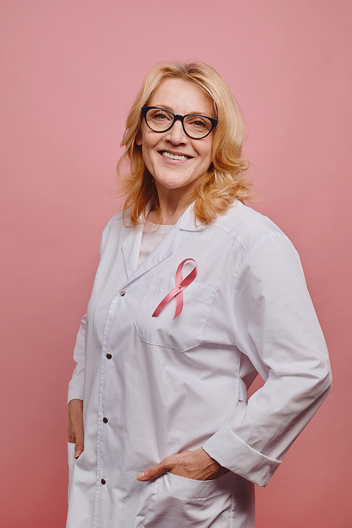 Vertical portrait of mature female doctor with pink ribbon on white coat smiling at camera while posing against pink background in studio