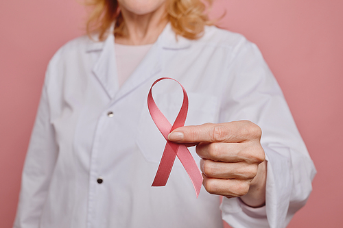 Close up of female doctor holding pink ribbon to camera while while posing against pink background in studio , breast cancer awareness concept, copy space