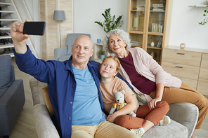 Portrait of modern senior couple taking selfie photo via smartphone with cute red haired girl in home interior, copy space