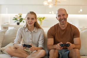 Warm-toned portrait of happy mature father playing video games with teenage daughter while sitting on couch in cozy home interior, copy space