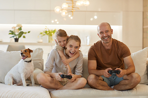 Cute warm-toned portrait of happy modern family playing video games together while sitting on couch in cozy home interior, smiling mature man enjoying time with two daughters and pet dog
