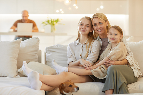 Warm-toned portrait of happy mother embracing two daughters while posing together sitting on couch in cozy home interior with father in background, copy space