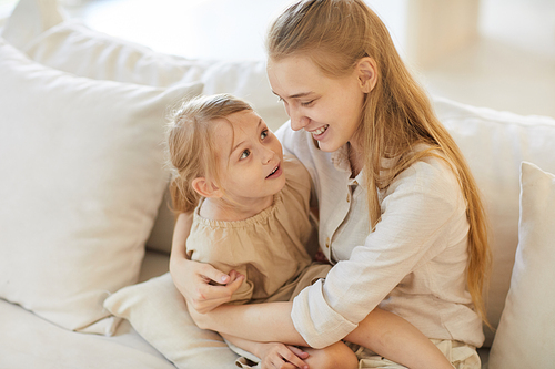 Warm toned high angle portrait of smiling teenage girl holding cute little sister while sitting on cozy white couch at home, copy space