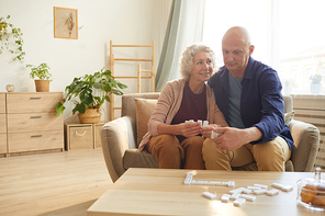 Warm toned portrait of happy senior couple holding domino pieces while playing board games at home, copy space