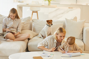 Warm-toned family scene of young mother helping little daughter draw or study with teenage girl and pet dog sitting on comfortable white couch in home interior, copy space