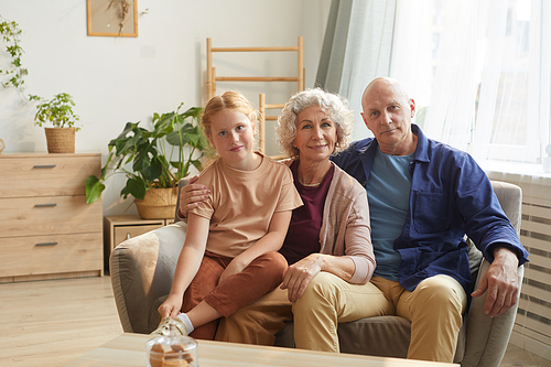 Warm toned portrait of happy senior couple posing with cute granddaughter while sitting on couch together in cozy home interior