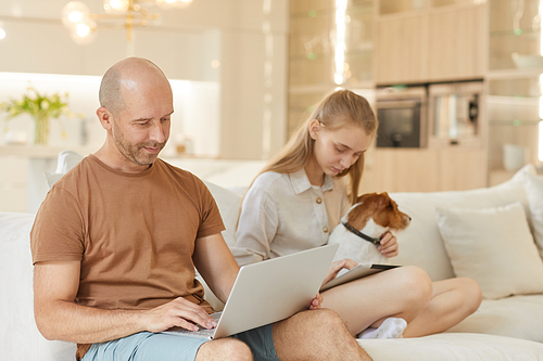 Warm-toned portrait of family using computers at home, focus on mature man holding laptop wile sitting on couch with teenage daughter, copy space