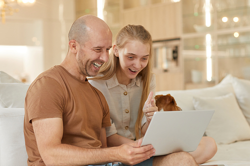 Warm-toned portrait of mature father smiling happily while looking at laptop screen with teenage girl while sitting on couch in home interior, copy space