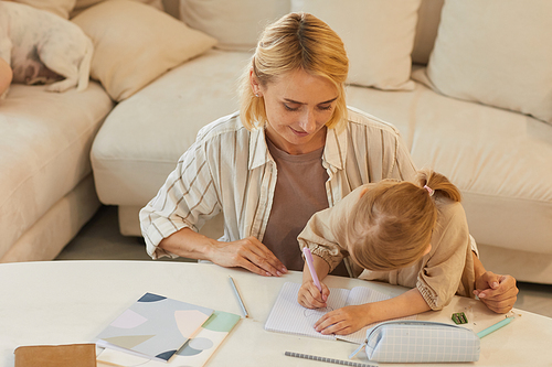 Warm-toned high angle portrait of happy young mother smiling while helping cute little girl drawing on studying at home, copy space