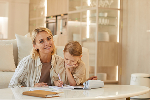 Warm-toned portrait of happy young mother smiling at camera while helping cute little girl drawing on studying at home, copy space