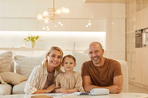 Warm-toned portrait of happy young family looking at camera and smiling while helping cute little girl drawing on studying at home, copy space