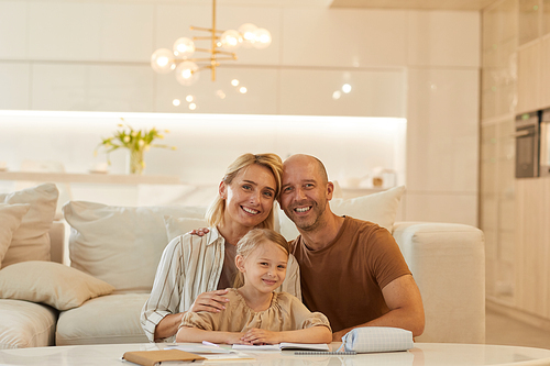 Warm-toned portrait of happy family looking at camera and smiling while helping cute little girl drawing on studying at home, copy space