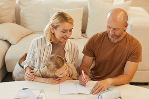 Warm-toned high angle portrait of happy family mother and father helping cute little girl drawing on studying at home, copy space