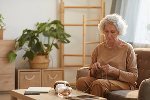 Warm toned portrait of content senior woman knitting in cozy home lit by sunlight, copy space