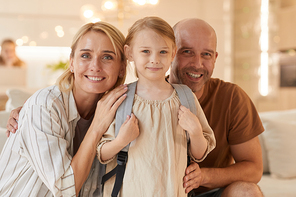 Warm-toned portrait of happy family looking at camera and smiling while posing with cute little girl carrying backpack