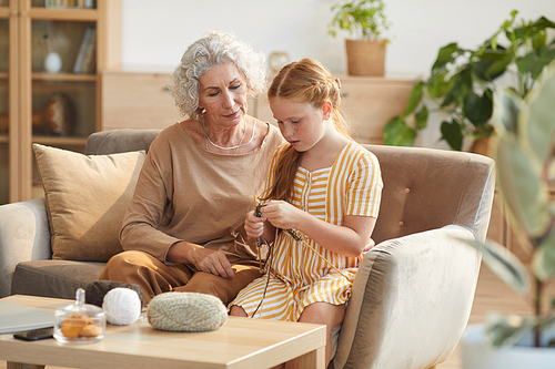 Warm toned portrait of elegant senior woman teaching granddaughter knitting while sitting on couch in cozy home lit by sunlight, copy space