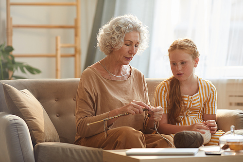 Warm toned portrait of elegant senior woman teaching granddaughter knitting while sitting on couch in cozy home lit by sunlight