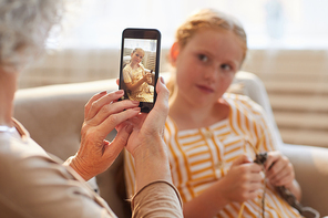 Warm toned portrait of unrecognizable senior woman taking smartphone photo of cute red haired girl in cozy home interior