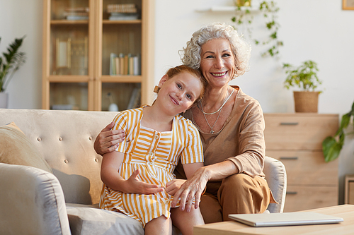 Warm toned portrait of smiling senior woman embracing cute granddaughter while sitting on couch and looking at camera in cozy home interior, copy space