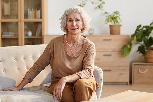 Warm toned portrait of elegant senior woman smiling at camera while sitting on couch and looking at camera in cozy home interior, copy space