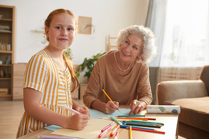 Warm toned portrait of cute red haired girl smiling cheerfully at camera while drawing together with grandma enjoying time at home, copy space