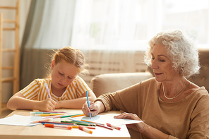 Warm toned portrait of smiling senior woman babysitting cute red haired girl and drawing together while sitting by coffee table in cozy living room