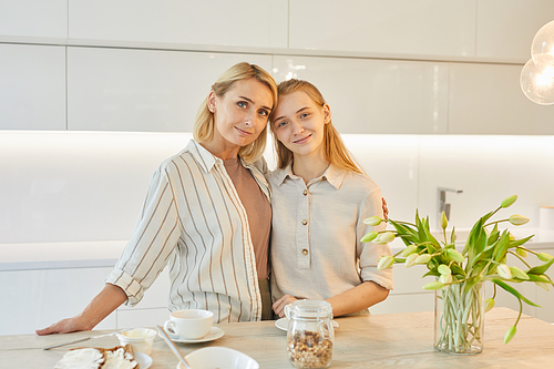 Warm-toned portrait of modern adult mother posing with teenage daughter in kitchen interior while enjoying breakfast together and looking at camera, copy space