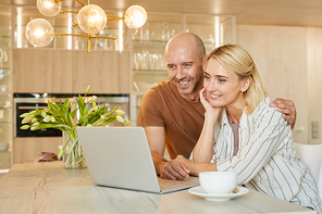 Warm-toned portrait of happy adult couple looking at laptop screen and smiling while talking by video chat with family, copy space