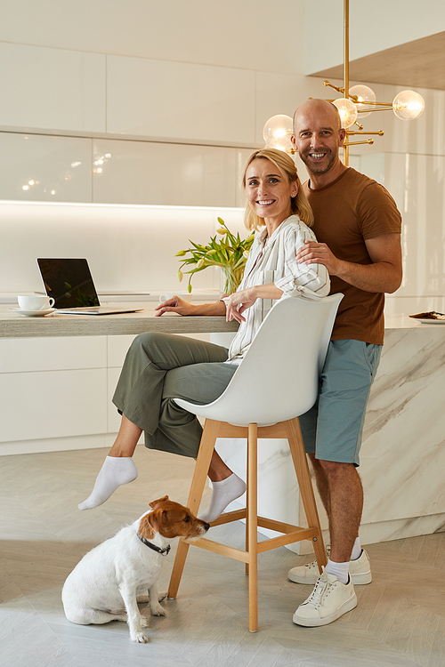 Warm-toned full length portrait of beautiful adult couple looking at camera while sitting at kitchen counter in minimal home interior, copy space