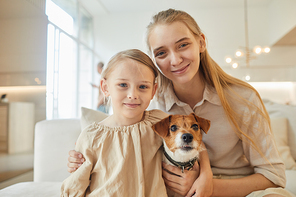 Warm-toned portrait of two sisters posing with pet dog and looking at camera while sitting on couch in minimal home interior, copy space