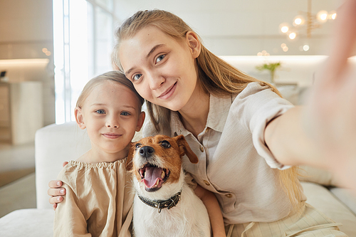Warm-toned portrait of two sisters taking selfie with pet dog and looking at camera while sitting on couch in minimal home interior, copy space