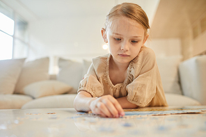 Warm-toned portrait of cute little girl solving puzzle alone while sitting on couch in minimal home interior, copy space
