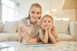 Warm-toned portrait of two sisters solving puzzle together while enjoying time indoors at home and looking at camera, copy space