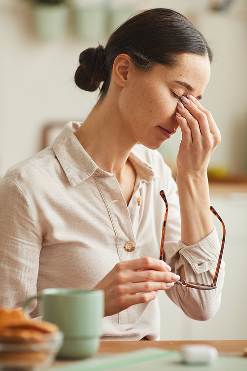 Vertical warm-toned portrait of tired young woman rubbing nose bridge and taking off glasses while suffering from headache