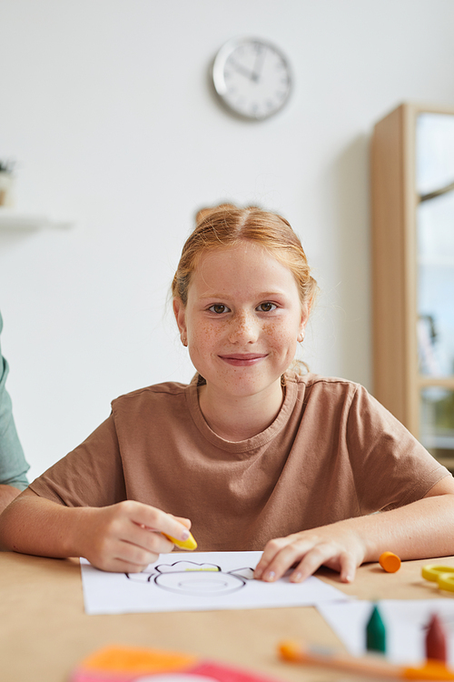 Vertical portrait of freckled red haired girl smiling at camera while drawing pictures with crayons during art class in school, copy space