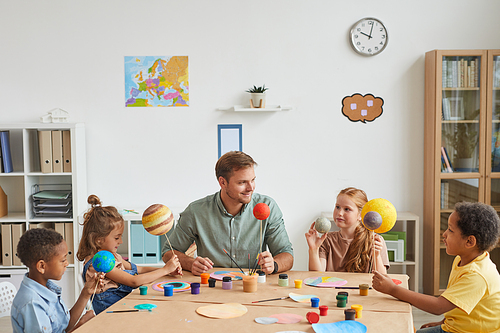 Portrait of smiling male teacher working with multi-ethnic group of children painting planet models in art and craft lesson, copy space