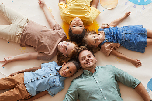 Above view at smiling male teacher with multi-ethnic group of children lying in circle and looking at camera while having fun in preschool or development center..