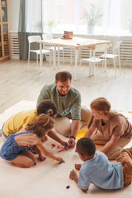 High angle portrait of young male teacher working with kids drawing pictures while having fun in preschool or development center, copy space