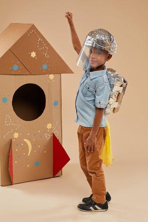 Vertical full length portrait of cute African-American boy playing astronaut in handcrafted space suit while standing against cardboard rocket in studio