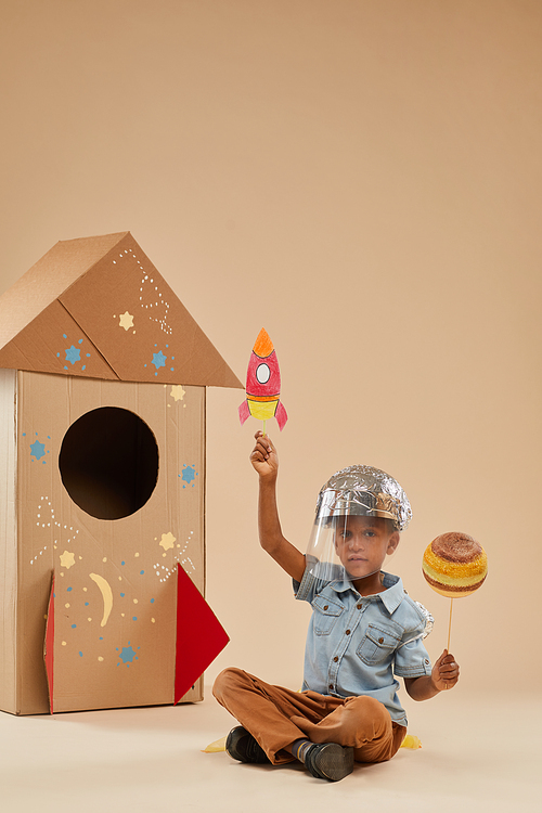 Vertical full length portrait of cute African-American boy playing astronaut in handcrafted space suit while sitting on floor against cardboard rocket in studio, copy space