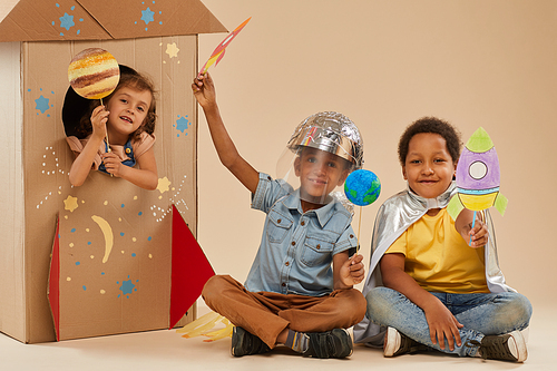 Full length portrait of cute children playing astronauts while sitting on floor against cardboard rocket in studioFull length portrait of cute children playing astronauts while sitting on floor against cardboard rocket in studio